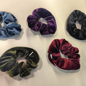 May include: Five velvet scrunchies in various colors: teal, purple, black, olive green, and burgundy. These hair accessories are made of a soft, textured fabric and are arranged on a light-colored surface.