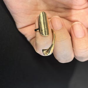 May include: A gold-toned metal nail ring featuring a smooth, elongated, shield-like plate that covers the fingernail area and a curved band that wraps around the base of the finger, creating a sleek, polished, and reflective surface finish.
