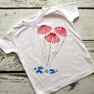 May include: White t-shirt with a red, white, and blue graphic of three parachutes. The parachutes are descending towards a gray rock with blue waves.