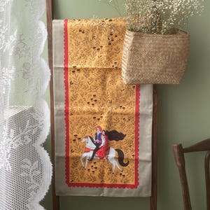 May include: A decorative textile with a mustard yellow floral pattern and a red scalloped border. The centre features an illustration of a couple riding a white horse. A woven basket with white flowers hangs above.