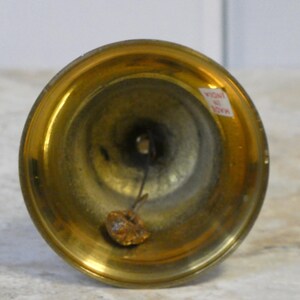 May include: A close-up of a small brass bell, showing its flared rim and rounded body. The bell has a warm gold colour. A small tag reading "MADE IN INDIA" is visible. The bell is likely a decorative item.