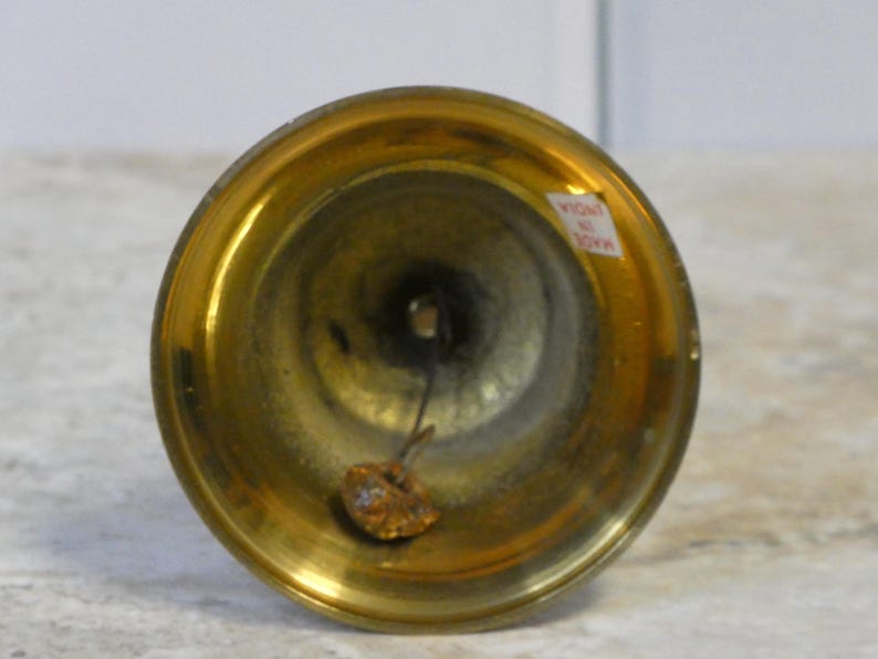 May include: A close-up of a small brass bell, showing its flared rim and rounded body. The bell has a warm gold colour. A small tag reading "MADE IN INDIA" is visible. The bell is likely a decorative item.