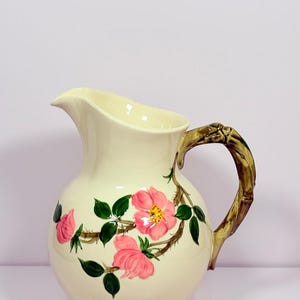 May include: A cream-colored ceramic pitcher featuring a hand-painted design of pink wild roses with green leaves and thorny brown stems. The handle is sculpted to resemble a textured, gnarled branch in shades of brown and green, adding detail.