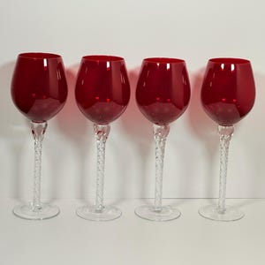 May include: Four red wine glasses with clear, twisted stems and round bases. The glasses have a deep red bowl and are arranged in a row against a white background. The stems have a textured, decorative design.