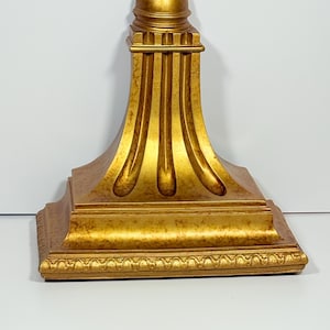 May include: A gold-colored decorative stand with a square base and a rounded top. The stand features vertical fluted detailing and ornate trim. The overall design is classical and elegant, suitable for displaying decorative items.