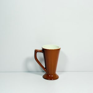 May include: A brown ceramic coffee mug with a handle. The mug has a flared shape, a pedestal base, and a white interior. The mug is set against a white background.