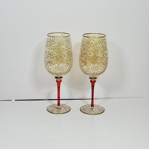 May include: Two decorative wine glasses with a gold filigree design on the bowl and rim. The stems are red with gold accents, and the base is gold with a matching filigree pattern. The glasses are for serving wine.