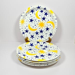 May include: A stack of four white ceramic plates with a celestial design. Each plate features a pattern of yellow crescent moons, yellow and blue stars, and small blue dots. The plates are stacked on top of each other.