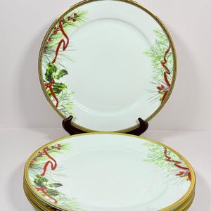 May include: A set of white porcelain dinner plates featuring a festive holiday pattern of green pine needles, red holly berries, and flowing red ribbons. Each plate is finished with a decorative gold rim and a thin black geometric border.