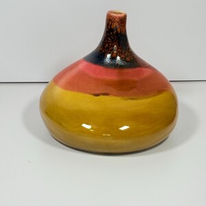 Peut inclure: Un vase en c&eacute;ramique avec une base arrondie et un col fin. Le vase est peint avec des bandes horizontales de jaune, d'orange, de noir et de marron. La finition brillante refl&egrave;te la lumi&egrave;re, soulignant les variations de couleur.