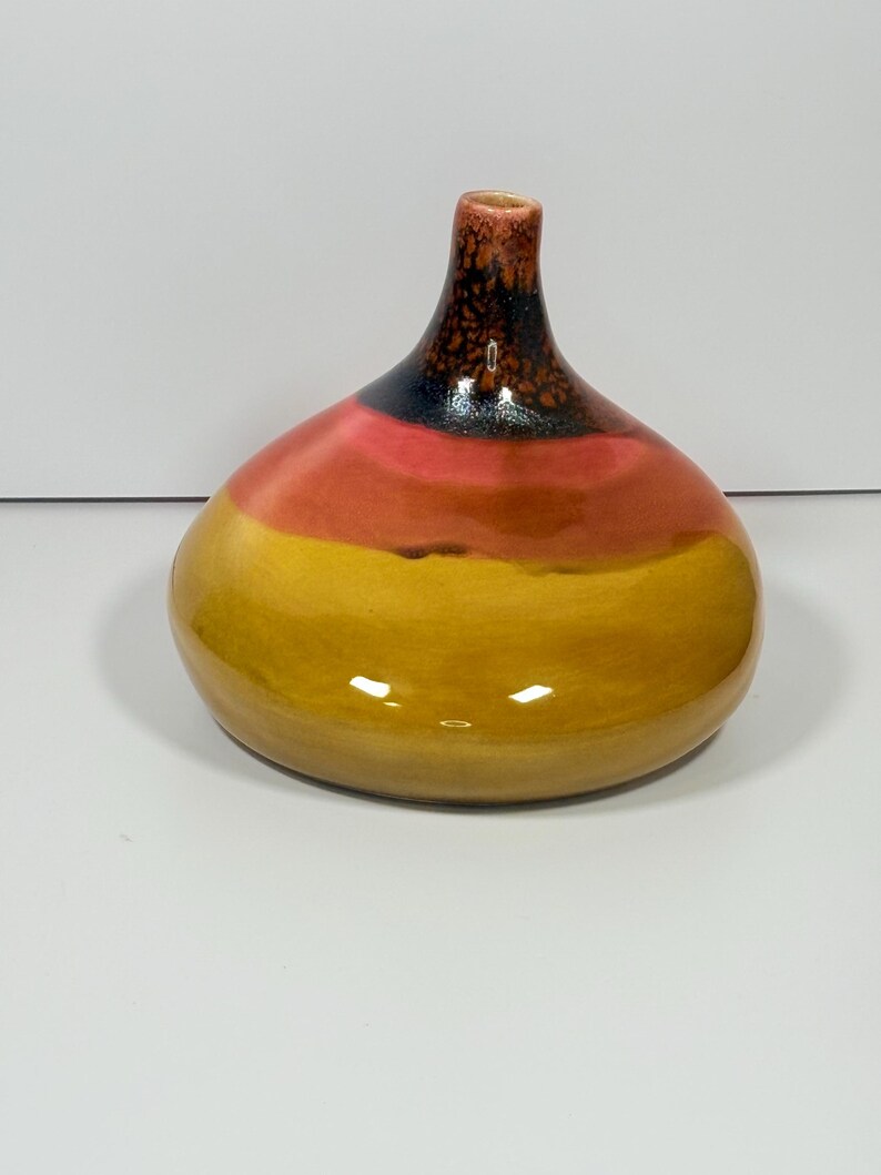 Peut inclure: Un vase en c&eacute;ramique avec une base arrondie et un col fin. Le vase est peint avec des bandes horizontales de jaune, d'orange, de noir et de marron. La finition brillante refl&egrave;te la lumi&egrave;re, soulignant les variations de couleur.