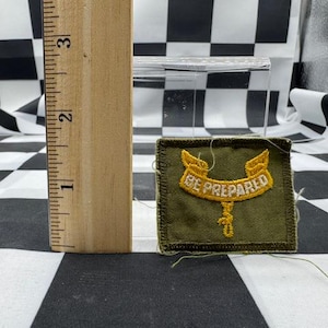 May include: A green and gold embroidered patch with the words "BE PREPARED". The patch is square and has a gold border. A wooden ruler is next to the patch, showing measurements in inches.