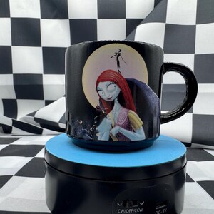 Puede incluir: Taza de cer&aacute;mica negra con Sally de Pesadilla antes de Navidad. La taza tiene un asa negra y un dise&ntilde;o de Sally frente a una luna llena. La taza est&aacute; sobre una plataforma azul.