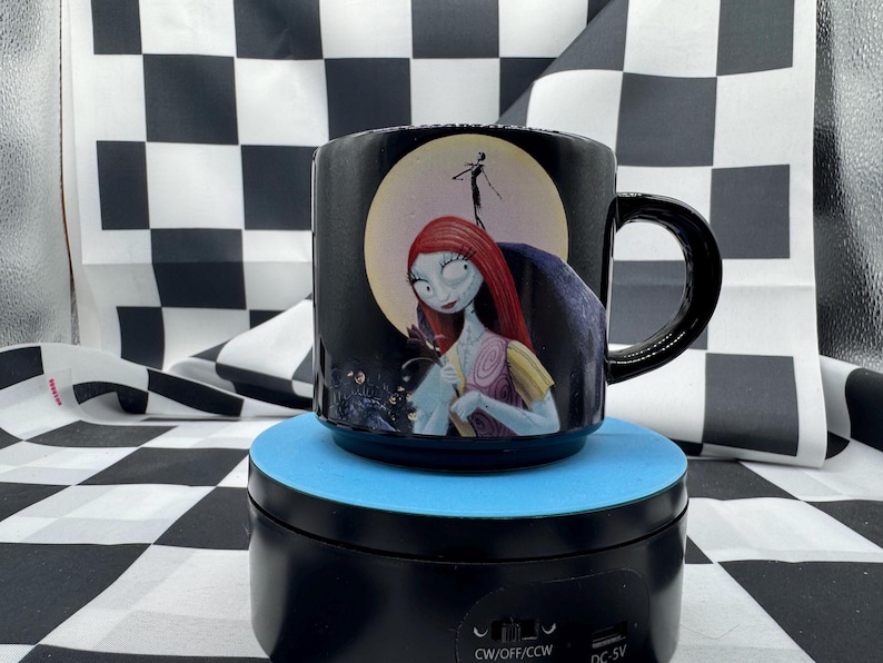 Puede incluir: Taza de cer&aacute;mica negra con Sally de Pesadilla antes de Navidad. La taza tiene un asa negra y un dise&ntilde;o de Sally frente a una luna llena. La taza est&aacute; sobre una plataforma azul.