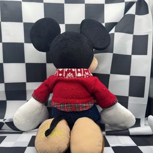 May include: A plush Mickey Mouse doll, viewed from the back, wearing a red sweater with a white and red patterned collar. The doll has black ears, a black tail, and tan feet. The background is a black and white checkered pattern.