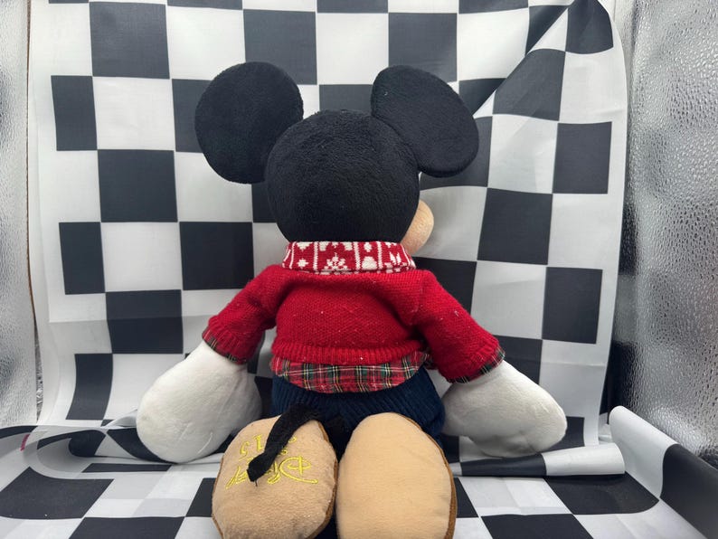 May include: A plush Mickey Mouse doll, viewed from the back, wearing a red sweater with a white and red patterned collar. The doll has black ears, a black tail, and tan feet. The background is a black and white checkered pattern.