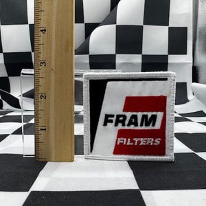 May include: A square patch with the text "FRAM FILTERS" in black and red, bordered in white. A wooden ruler is next to the patch, showing measurements in inches. The background features a black and white checkered pattern.