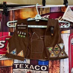 May include: Brown Girl Scout vest with patches and the text "GIRL SCOUTS USA SAN GORGONO 1349". The vest has various triangular patches with different designs and colors, hanging on a white hanger.