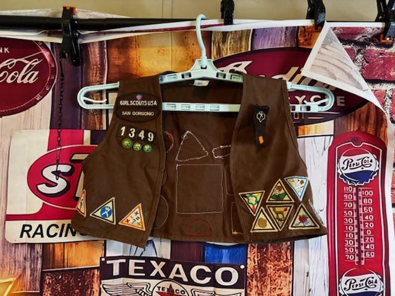 May include: Brown Girl Scout vest with patches and the text "GIRL SCOUTS USA SAN GORGONO 1349". The vest has various triangular patches with different designs and colors, hanging on a white hanger.