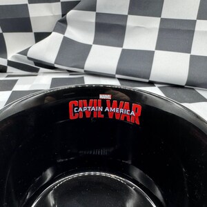 Puede incluir: Cuenco negro con el texto "CAPTAIN AMERICA CIVIL WAR" en rojo y blanco. El cuenco tiene un borde plateado y est&aacute; sobre un fondo a cuadros blanco y negro. Tambi&eacute;n es visible el logotipo de Marvel.