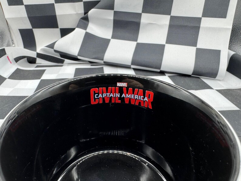 Puede incluir: Cuenco negro con el texto "CAPTAIN AMERICA CIVIL WAR" en rojo y blanco. El cuenco tiene un borde plateado y est&aacute; sobre un fondo a cuadros blanco y negro. Tambi&eacute;n es visible el logotipo de Marvel.