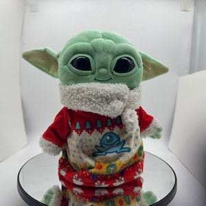 May include: A plush Grogu doll, or Baby Yoda, in a red and white festive sweater with a frog design. The doll has large black eyes, green skin, and large ears. The sweater features a white, fluffy collar.