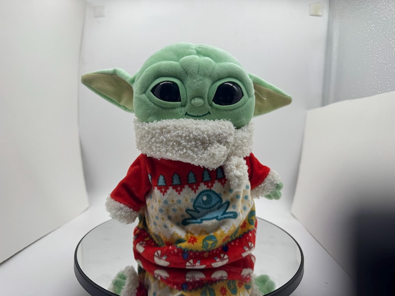 May include: A plush Grogu doll, or Baby Yoda, in a red and white festive sweater with a frog design. The doll has large black eyes, green skin, and large ears. The sweater features a white, fluffy collar.