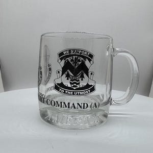 May include: A clear glass mug featuring a black printed emblem with the text WE SUPPORT TO THE UTMOST, SUPPORT COMMAND (A), and AMERICA. The design includes a central shield graphic with a letter M, pine trees, and a circular base element.
