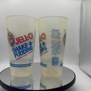 May include: Two clear plastic Jell-O Shake-A-Pudding shakers. The shakers have blue and red text and graphics. The text includes "JELL-O", "SHAKE-A-PUDDING", and "INSTANT PUDDING SHAKER". One shaker has directions printed on it.