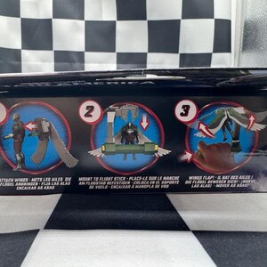 May include: A product box with instructions for attaching wings to a figure. The box features three circular illustrations with text in multiple languages. The background is a black and white checkered pattern.