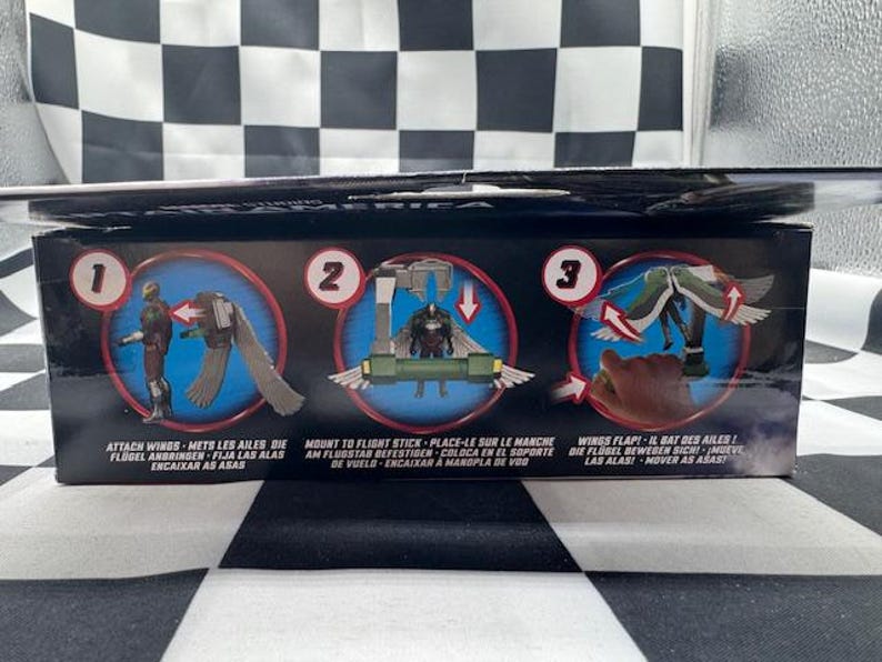 May include: A product box with instructions for attaching wings to a figure. The box features three circular illustrations with text in multiple languages. The background is a black and white checkered pattern.