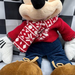May include: A plush Mickey Mouse doll wearing a red sweater and a white and red snowflake-patterned scarf. The doll has blue pants, brown shoes, and white gloves with black lines. The background is a black and white checkered pattern.