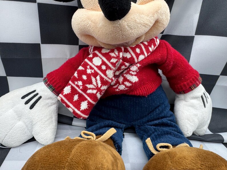 May include: A plush Mickey Mouse doll wearing a red sweater and a white and red snowflake-patterned scarf. The doll has blue pants, brown shoes, and white gloves with black lines. The background is a black and white checkered pattern.