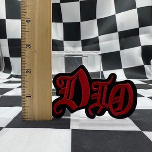 May include: Black and red embroidered patch with the word "DIO" in a gothic font. The patch is approximately 3 inches long and is displayed against a black and white checkered background.