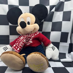 May include: A plush Mickey Mouse doll wearing a red sweater and a patterned scarf. The doll has black ears, a tan face, and white gloves. The doll's feet are brown, and the year "2015" is printed on the bottom of one foot.