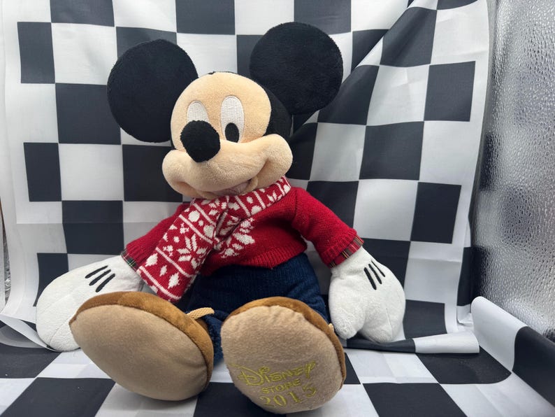 May include: A plush Mickey Mouse doll wearing a red sweater and a patterned scarf. The doll has black ears, a tan face, and white gloves. The doll's feet are brown, and the year "2015" is printed on the bottom of one foot.