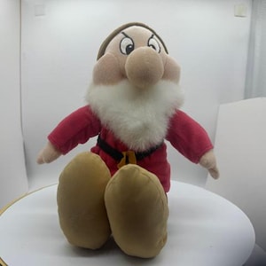May include: A plush toy of the character Grumpy from Snow White and the Seven Dwarfs. The figure features a brown hat, a red tunic with a black belt, tan shoes, and a thick white faux fur beard. The face has a frowning expression.
