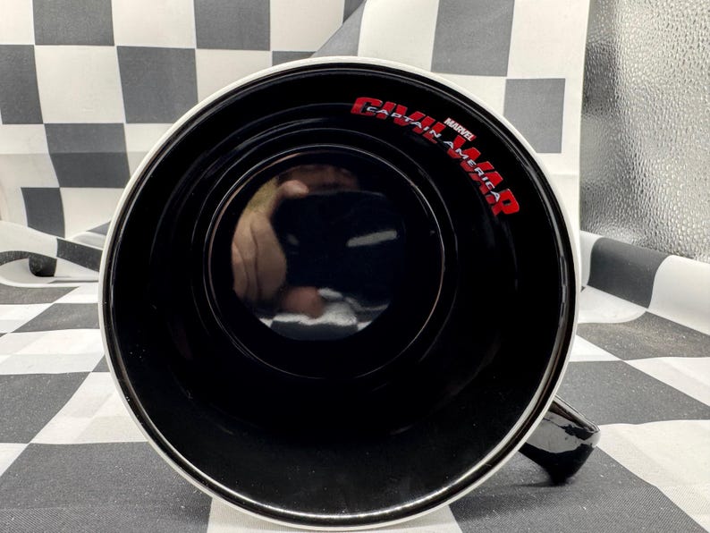 Puede incluir: Taza de cer&aacute;mica negra con borde y asa blancos. El interior es negro y las palabras "CAPTAIN AMERICA CIVIL WAR" est&aacute;n impresas en rojo. La taza est&aacute; sobre una superficie a cuadros blancos y negros.