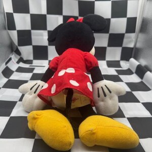 May include: A plush Minnie Mouse doll, viewed from the back, with a red dress with white polka dots, black ears, and a red bow. The doll has white gloves and yellow shoes. The background is a black and white checkered pattern.