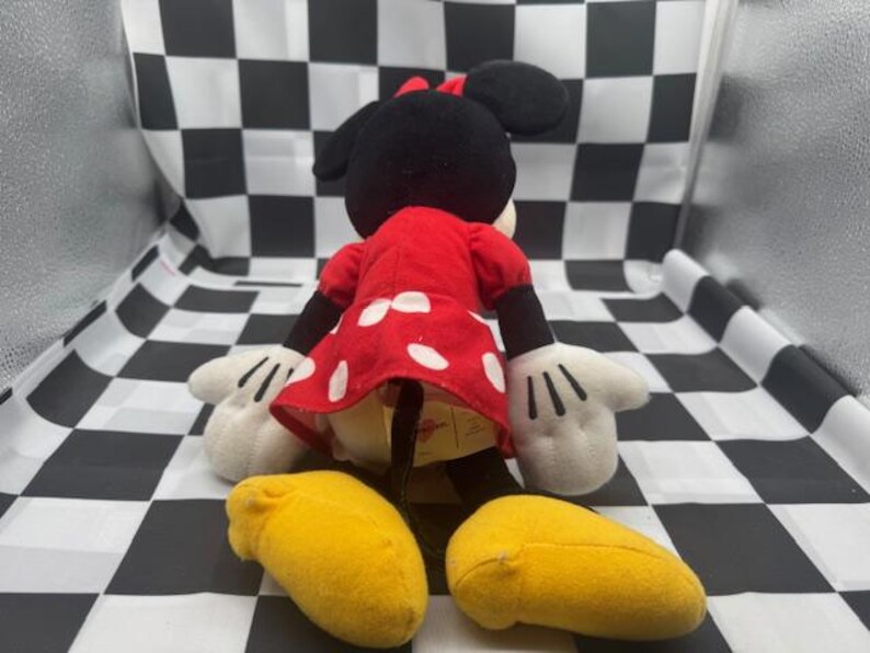 May include: A plush Minnie Mouse doll, viewed from the back, with a red dress with white polka dots, black ears, and a red bow. The doll has white gloves and yellow shoes. The background is a black and white checkered pattern.