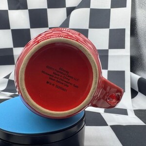 Puede incluir: Una taza de cer&aacute;mica roja con asa, vista desde abajo. La taza tiene un patr&oacute;n texturizado y un interior rojo. La base de la taza tiene texto impreso, incluyendo "&copy;Disney" y "Made in China".