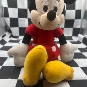 May include: A plush Minnie Mouse doll, featuring a red dress with white polka dots, black arms, and white gloves. The doll has yellow shoes and a black bow on her head. The background is a black and white checkered pattern.