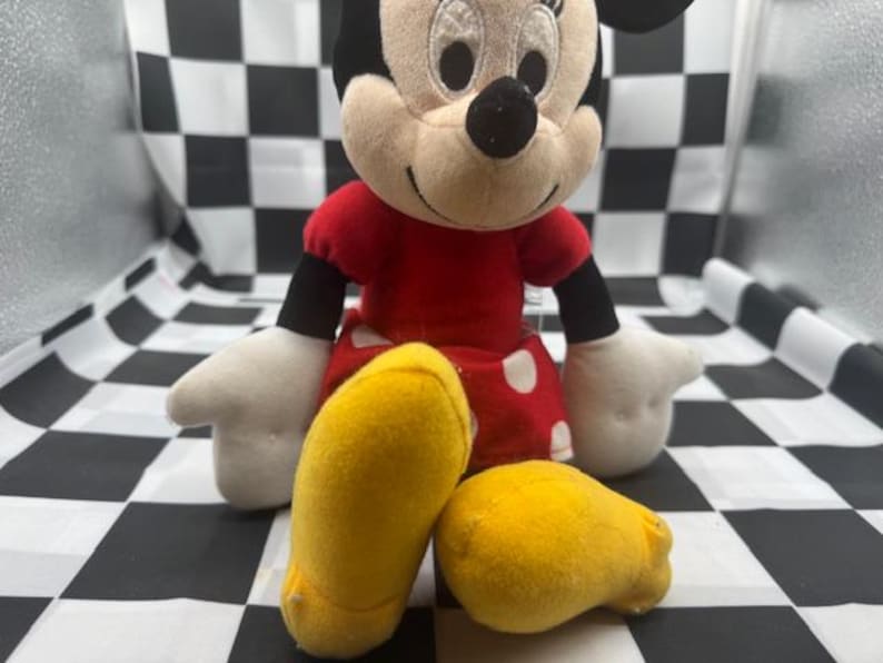 May include: A plush Minnie Mouse doll, featuring a red dress with white polka dots, black arms, and white gloves. The doll has yellow shoes and a black bow on her head. The background is a black and white checkered pattern.