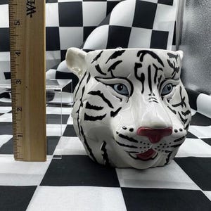 Peut inclure: Pot de fleurs en céramique blanche en forme de tête de tigre avec des rayures noires, des yeux bleus et un nez rose. Le pot mesure environ 7.6 cm de haut. Le fond présente un motif à carreaux noir et blanc.