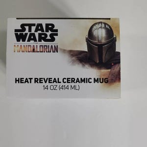 Pode incluir: Uma caixa branca com o logotipo de Star Wars: The Mandalorian e uma representa&ccedil;&atilde;o do capacete do Mandaloriano. A caixa diz "Heat Reveal Ceramic Mug" e tem uma capacidade de 414 ml. O design tem um tema espacial.