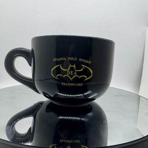 May include: A black ceramic coffee mug with a curved handle. The mug features gold text and a bat symbol, with the words "ATLANTA FIELD DIVISION DEA TRAINING UNIT". The mug is sitting on a reflective surface.