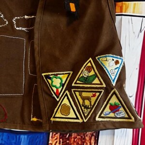 May include: Brown fabric with embroidered patches. The patches are triangular with yellow borders and feature various designs, including a deer, a chef, and a tomato. The fabric has white stitching and a small orange square.