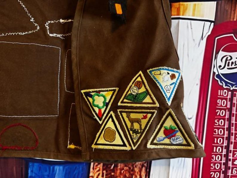 May include: Brown fabric with embroidered patches. The patches are triangular with yellow borders and feature various designs, including a deer, a chef, and a tomato. The fabric has white stitching and a small orange square.