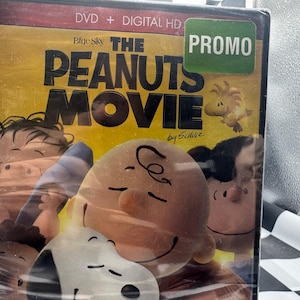 May include: The Peanuts Movie DVD + Digital HD. The DVD case features a yellow background with the movie title in large black letters. The cover art includes illustrations of Charlie Brown, Snoopy, and other Peanuts characters. A green "PROMO" sticker is in the upper right corner.