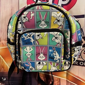 May include: A colorful Bugs Bunny mini backpack with a repeating comic strip design. The backpack features a front pocket and black straps. The comic strip squares have Bugs Bunny in various poses with speech bubbles and phrases.